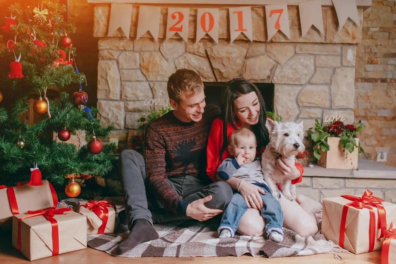 A family sitting in front of the fireplace with their baby, their dog, and a Christmas tree.